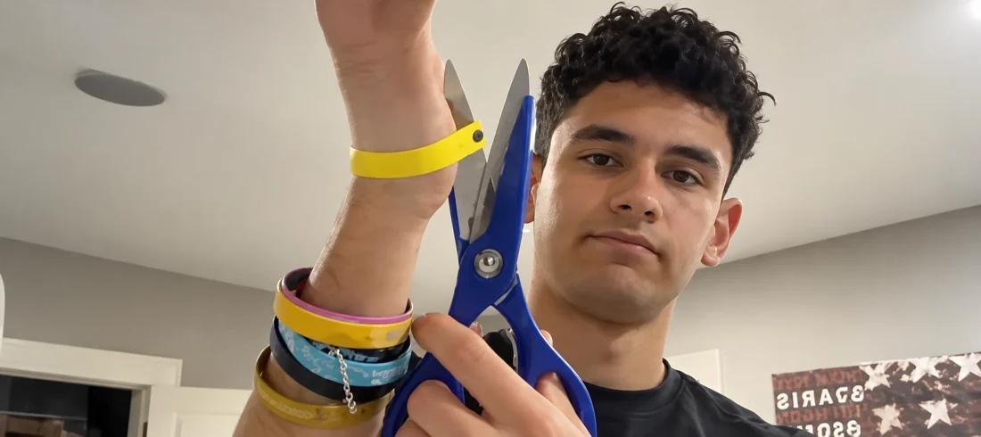 A young man holds scissors beside a yellow wristband as he prepares to remove it.